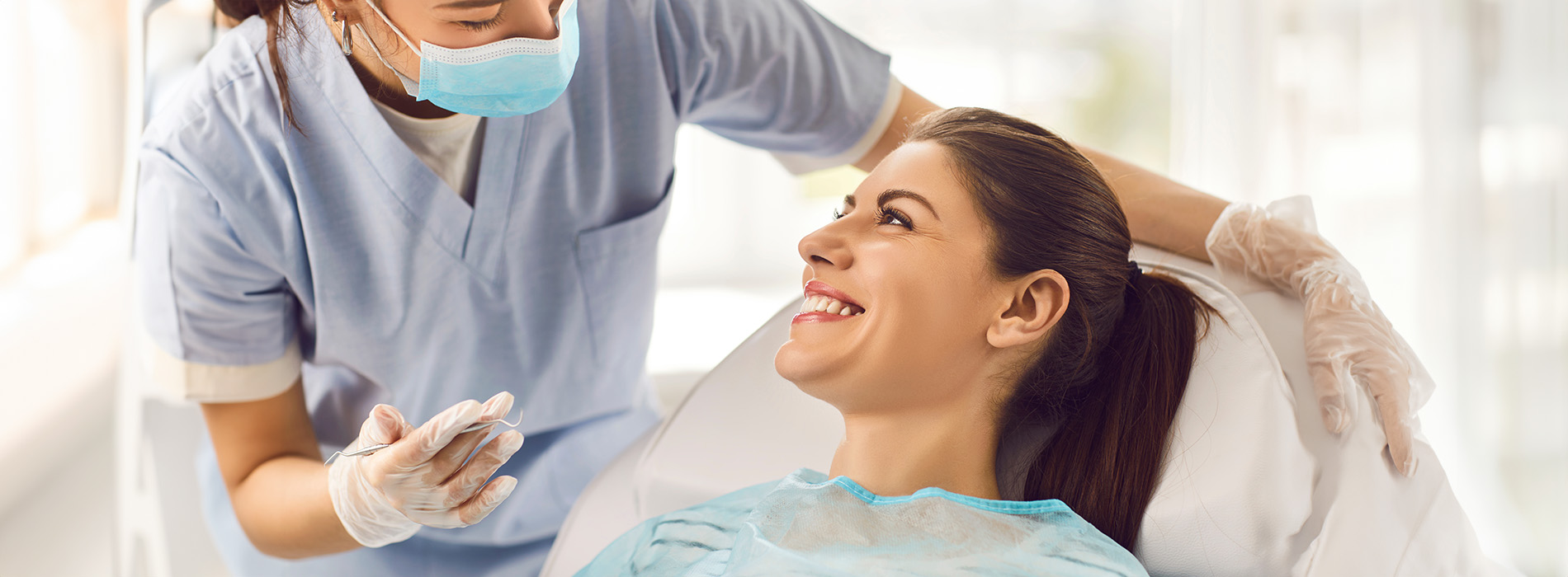 A person is seated in a dental chair, receiving care from a dental professional who stands behind them.